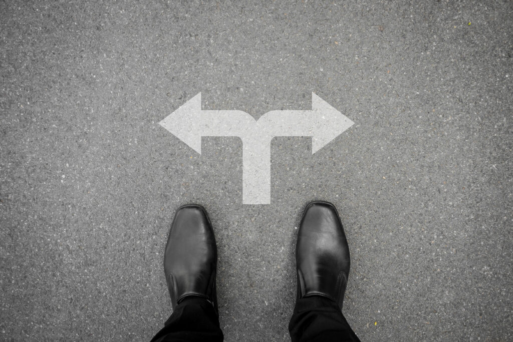 Person standing on pavement at a fork in the road with white arrows painted on the ground pointing left and right