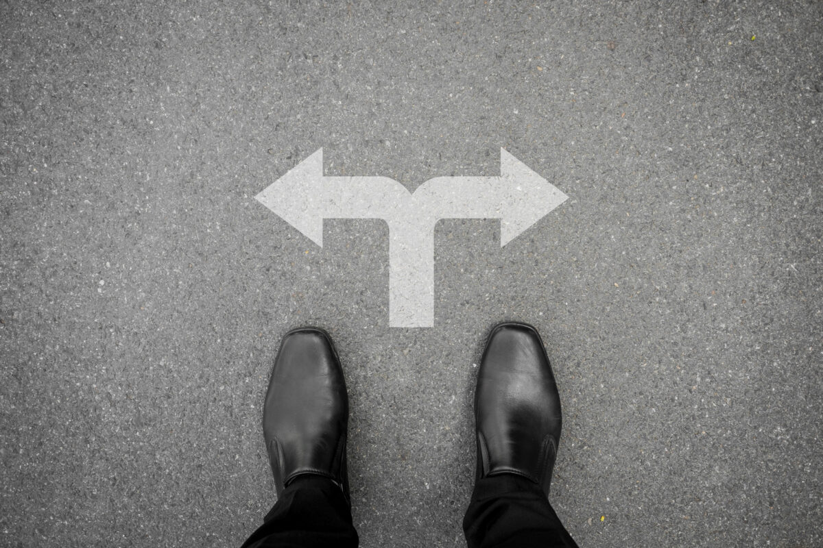 Person standing on pavement at a fork in the road with white arrows painted on the ground pointing left and right