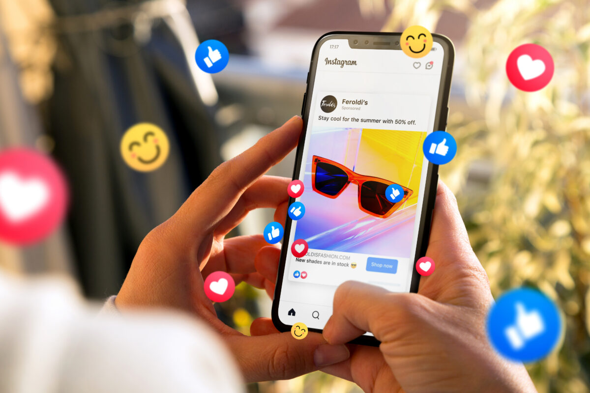 A person holding a smartphone showing an Instagram shopping ad for sunglasses, surrounded by floating like, heart, and smile reaction icons representing social engagement.